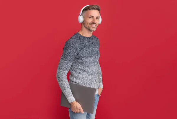 Happy man in casual listen to audio course in headphones holding laptop red background, e-learning — Stock Photo, Image