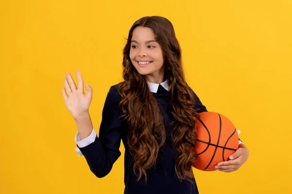 Felice teen girl tenere palla da basket agitando ciao su sfondo giallo, scuola di sport — Foto Stock