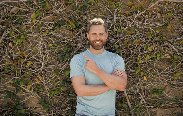 Счастливый бородатый взрослый мужчина в летней рубашке указывая пальцем на естественный фон, реклама — стоковое фото