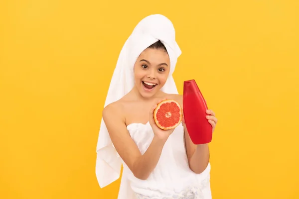 Verbaasd tiener kind in handdoek met grapefruit shampoo fles op gele achtergrond — Stockfoto