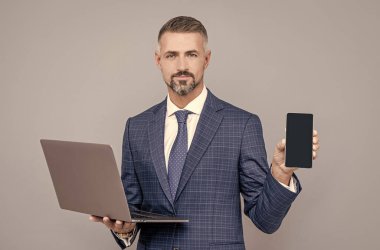 Takım elbiseli kendine güvenen işadamı dizüstü bilgisayar tutuyor ve akıllı telefon ve reklam sunuyor.