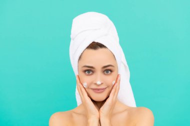 Gülümseyen genç bir kadın mavi arka plana yüz kremi sürüyor.