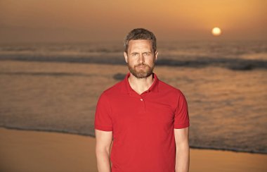 Yaz tişörtlü sakallı yakışıklı adam sahilde gün batımlı, yaz tatilinde.