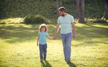 Mutlu oğul ve baba, güneşli bir yaz gününde parkta, çimlerde, ailede el ele tutuşuyorlar.
