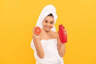 Sarı arka planda greyfurt şampuanı şişesiyle havluyla gülümseyen çocuk.