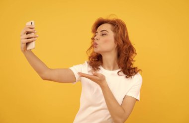 Kadın cep telefonuna el öpme hareketi yapıyor. Sarı arka plan, selfie.