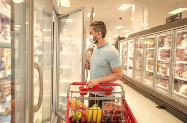 Alışveriş arabalı, koruyucu maskeli bir adam marketten yiyecek alıyor, alışveriş yapıyor.