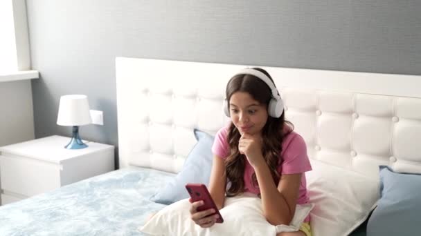 Niña feliz hablando en cámara de teléfono inteligente en auriculares para bloguear en el dormitorio, la vida moderna — Vídeo de stock