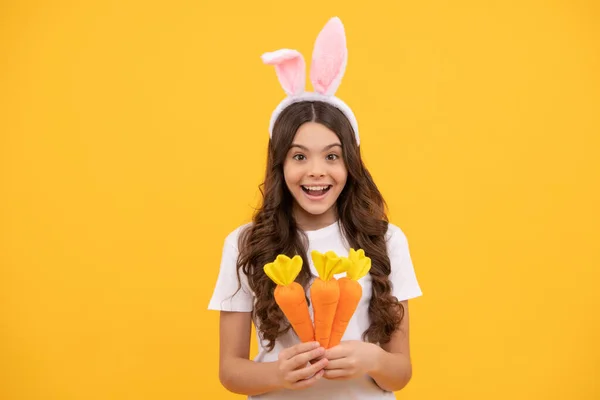 Zaskoczone dziecko w uszach króliczka trzymać marchewkę na żółtym tle, Wielkanoc — Zdjęcie stockowe