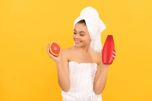 Vrolijk tiener kind in handdoek met grapefruit shampoo fles op gele achtergrond — Stockfoto