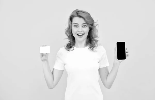 Chica sorprendida mostrando tarjeta de crédito o débito y teléfono inteligente para hacer compras en línea, compras en línea. —  Fotos de Stock