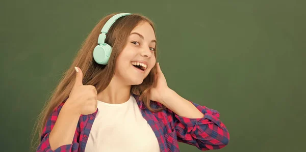 Fille heureuse tenir écouteurs modernes au lycée au tableau noir montrant pouce vers le haut, accessoire audio. — Photo