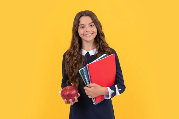 Wesoły dziecko trzymać szkolne książki do pracy domowej i lunch jabłko na żółtym tle, obiad — Zdjęcie stockowe