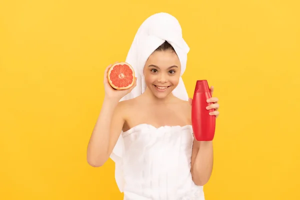 Glücklich Teenager-Kind im Handtuch mit Grapefruit-Shampoo-Flasche auf gelbem Hintergrund — Stockfoto