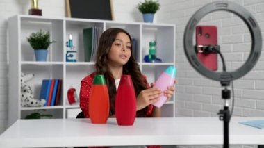 Uzun saçlı bir çocuk şampuan şişelerinde selfie lambası ve telefon ve güzellik bloguyla video eleştirisi yapıyor.