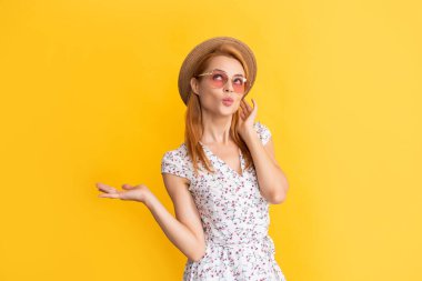 Sarı arka planda, hasır şapkalı ve güneş gözlüklü gülümseyen genç kadın.