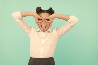 Mutlu okul çağında çocuk parmaklarını süper kahraman maskesi yapar dilini mavi arka plana sokar, oynar