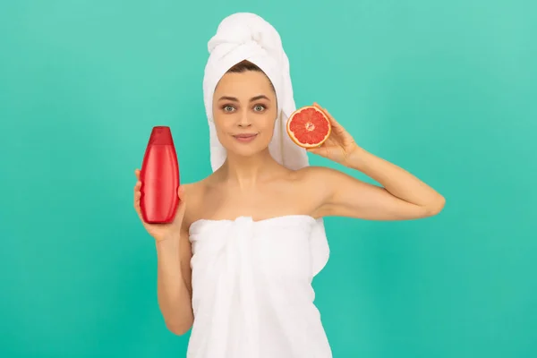 Smiling young woman in towel with grapefruit shampoo bottle on blue background — Fotografia de Stock