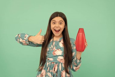 Şaşırmış uzun saçlı çocuk şampuan şişesini tut baş parmağını kaldır, cilt bakımı