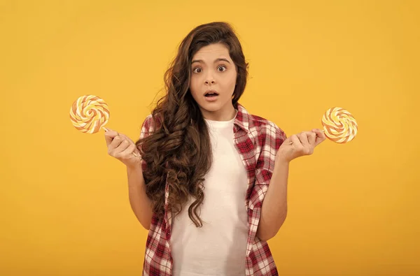 Geschokt tiener meisje met krullend haar houden lolly karamel snoep op gele achtergrond, zoetwaren. — Stockfoto