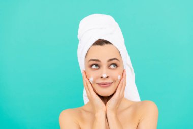 Neşeli genç kız mavi arka plana yüz kremi sürüyor.