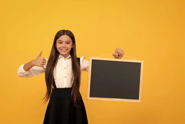 Menina adolescente alegre em uniforme segurar escola quadro negro para espaço de cópia mostrando polegar para cima, apresentando — Fotografia de Stock