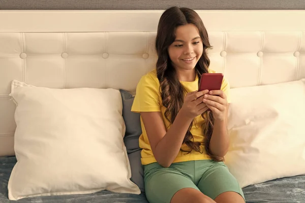 Happy kid texting on mobile phone while sitting in bed, text — Stockfoto