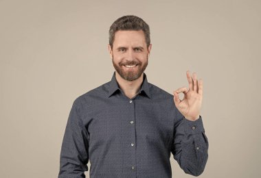 Yüzü sakallı mutlu bir adam. Sıradan bir tişörtle gülümsüyor. Yüzük jestleri gri arka plan gösteriyor.