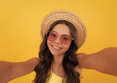 Çocuk hasır şapka ve gözlük takıyor. Sarı arka planda kıvırcık saçlı neşeli genç kız..