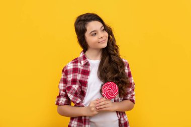 Uzun kıvırcık saçlı gülümseyen çocuk sarı arka planda lolipop karamel şekeri tutuyor, şeker düşkünü..
