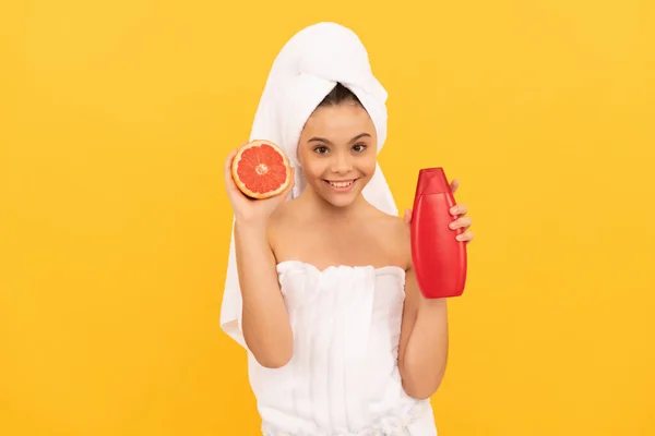 Vrolijk meisje in handdoek met grapefruit shampoo fles op gele achtergrond — Stockfoto