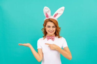 Tavşan kulaklı ve papyonlu mutlu Paskalya kadını mavi arka planda ürünü ve işaret parmağını sunar.