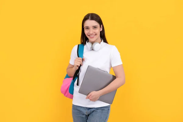 Young girl with backpack hold laptop in headphones on yellow background, education — Stockfoto