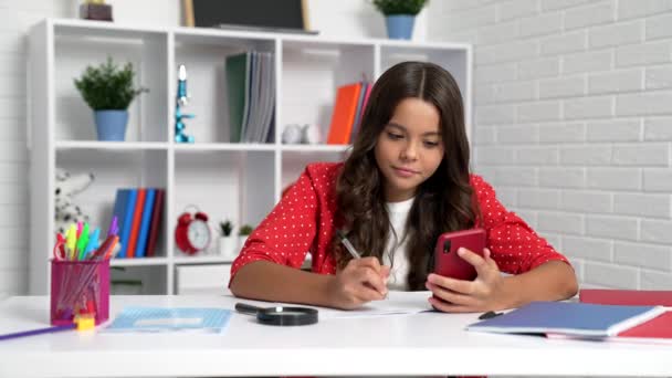 Серйозна дівчинка робить домашнє завдання за допомогою мобільного телефону на шкільному столі, навчання смартфона — стокове відео