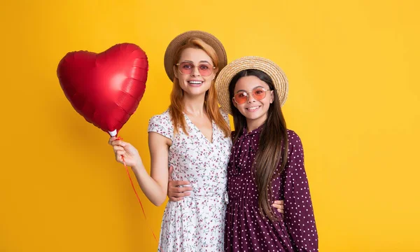 Positive mother and daughter hold love heart balloon on yellow background — Zdjęcie stockowe