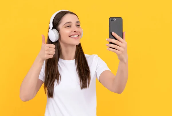 Smiling woman listen music in earphones with smartphone show thumb up on yellow background, selfie — Photo