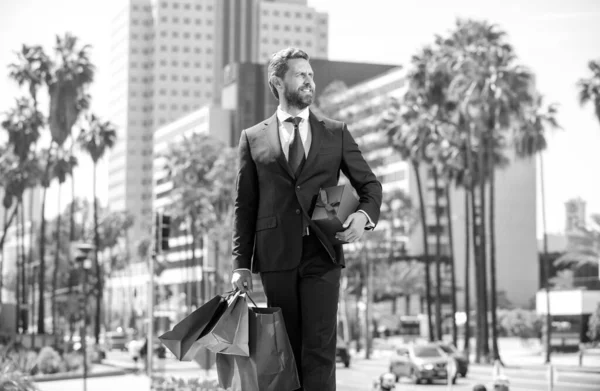 Hombre seguro de sí mismo en traje elegante caminar con bolsas de compras y caja de regalos en el exterior, compra — Foto de Stock