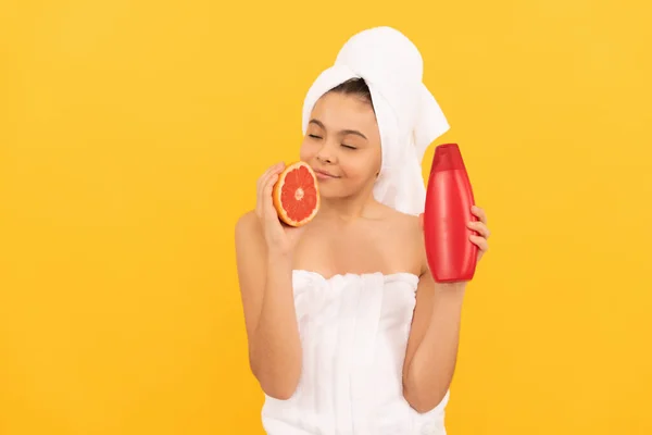 Smiling teen girl in towel with grapefruit shampoo bottle on yellow background — Stock Photo, Image