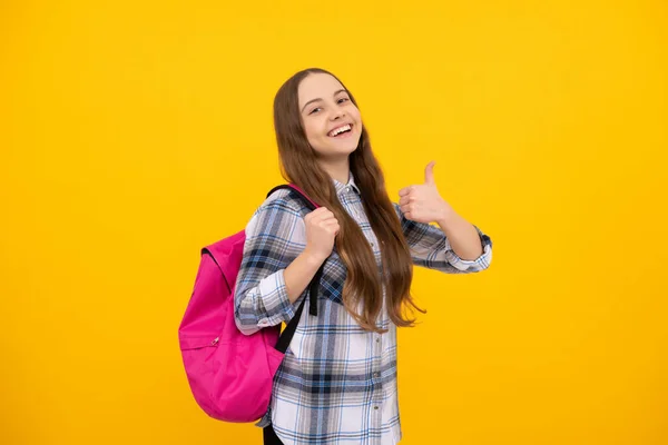 Χαρούμενη έφηβη κοπέλα με καρό πουκάμισο φέρει σακίδιο που δείχνει τον αντίχειρα επάνω στο κίτρινο φόντο, σχολείο — Φωτογραφία Αρχείου