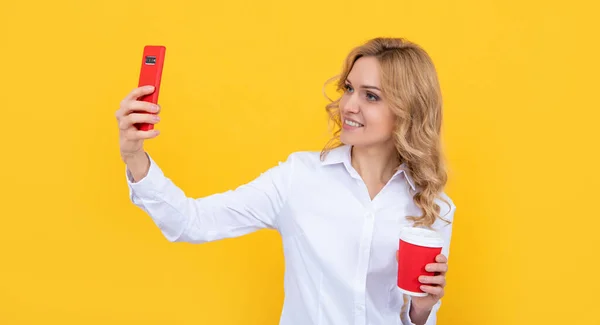 Glad blond kvinna med kaffekopp gör selfie på telefon på gul bakgrund — Stockfoto