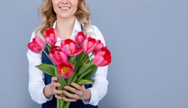 glad young woman in apron with spring tulip flowers on grey background