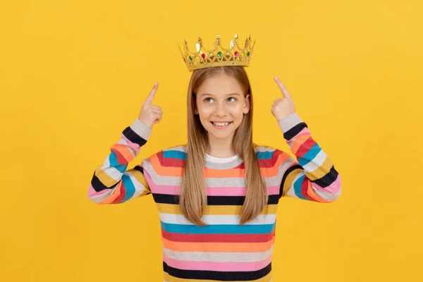 행복 한 십 대 아이가 노란 배경에 있는 퀸 왕관을 손가락으로 가리키는 모습 — 스톡 사진