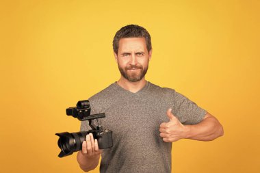 Neşeli sakallı adam kameramanla video bloğu için film çekiyor, başparmağını gösteriyor, filme çekiyor.