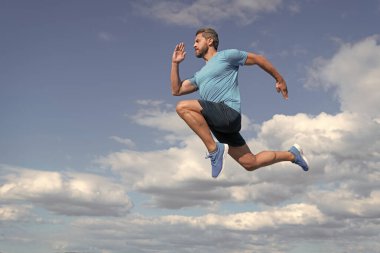 Kaslı vücutlu enerjik erkek atlet gökyüzü arka planında spor kıyafetleri ve dayanıklılıkla koşuyor.