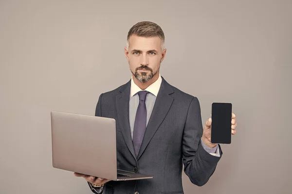 Confident businessman man in businesslike suit hold computer and presenting smartphone, advert — Stock Photo, Image