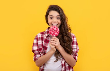 Uzun kıvırcık saçlı mutlu çocuk sarı arka planda lolipop karamel şekeri yaladı, çocukluk..