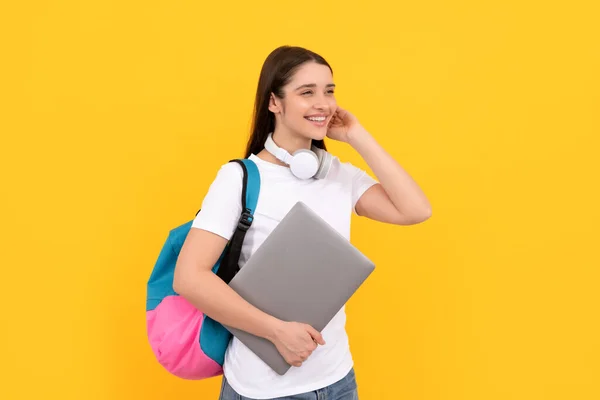 Wesoła młoda dziewczyna z plecakiem trzymać laptopa w słuchawkach na żółtym tle, e-learning — Zdjęcie stockowe