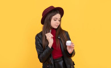 Sarı arka planda makyaj fırçası ve pudra tutan şapkalı pozitif çocuk, moda.