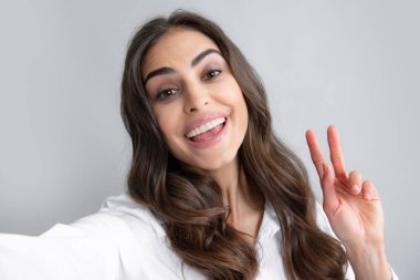 Genç ve güzel bir kadının selfie çekerken yakın çekimi. İzole edilmiş gri arka plan. Barış işaretiyle poz veren güzel kız.