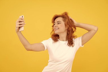 Heyecanlı, mutlu genç bir kadının görüntüsü, sarı arka planda izole edilmiş, kamerayla selfie çekiliyor..